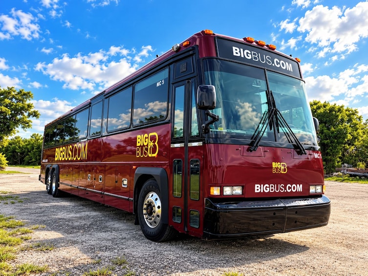 A red double-decker bus with 'BIGBUS.COM' branding parked on a gravel surface. 2