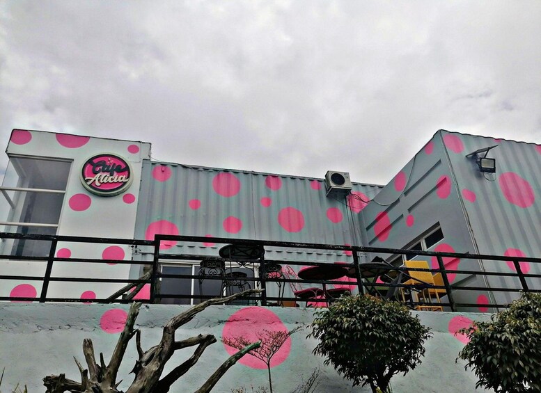 Ein Gebäude mit einem pinkfarbenen und grauen Punkte-Muster, einer Schrifttafel mit der Aufschrift 'Café Alicia' und Außenbestuhlung mit Tischen und Stühlen. 2