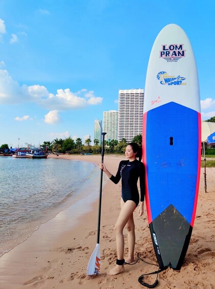 Eine Person steht auf einem Sandstrand und hält ein Stand-Up-Paddleboard, im Hintergrund sind Stadtansicht und Boote zu sehen. 3