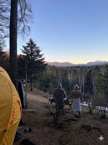 To personer går i et skovområde med et telt og et bjergrigt landskab i baggrunden. 1
