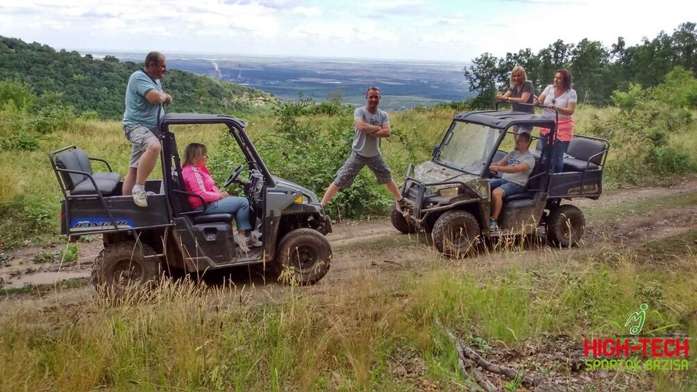 Two off-road vehicles on a dirt path with people on them, surrounded by greenery and a distant view of a landscape. 5