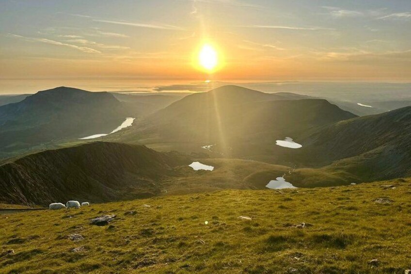 A mountainous landscape at sunrise with the sun low on the horizon, casting long shadows. 2