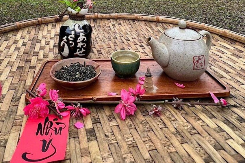 A traditional tea set on a woven tray with a teapot, a cup, and a bowl of tea leaves, accompanied by pink flowers and a red tag. 4