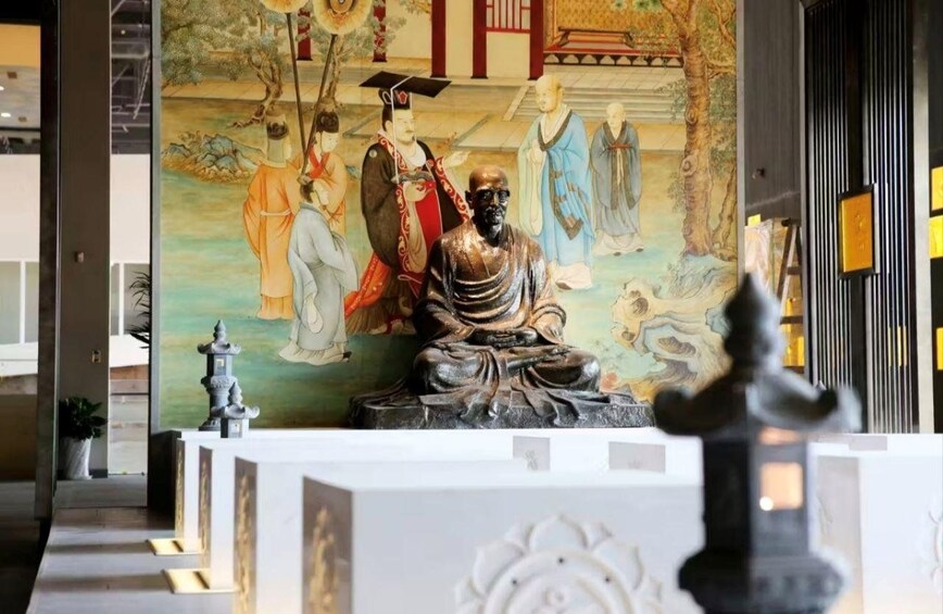 Une statue représentant une figure assise, vêtue d’une longue robe, avec une peinture chinoise traditionnelle en arrière-plan et la façade d’un bâtiment moderne. 2