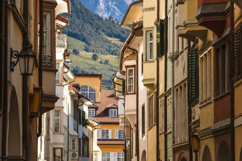 A narrow street flanked by historic buildings with balconies and shutters, leading towards a mountainous backdrop. 4