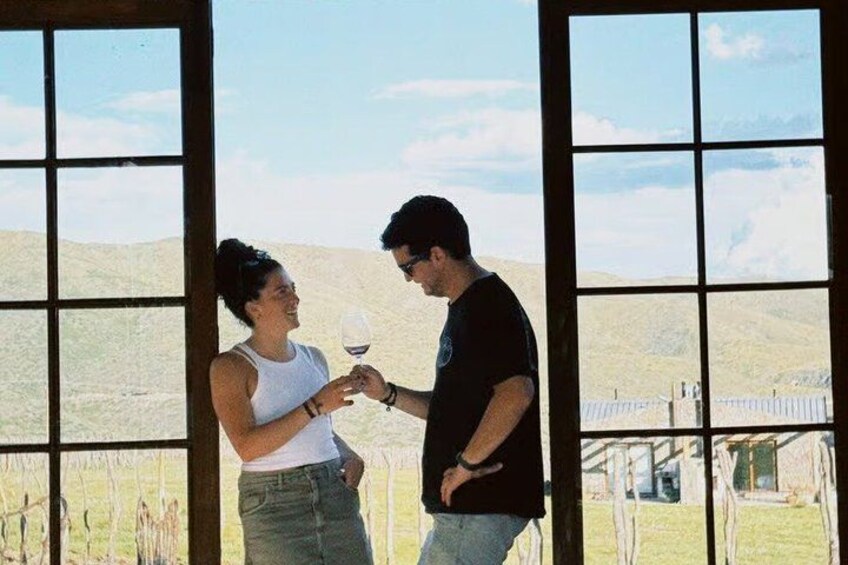 Two people standing indoors with a vineyard in the background. 1