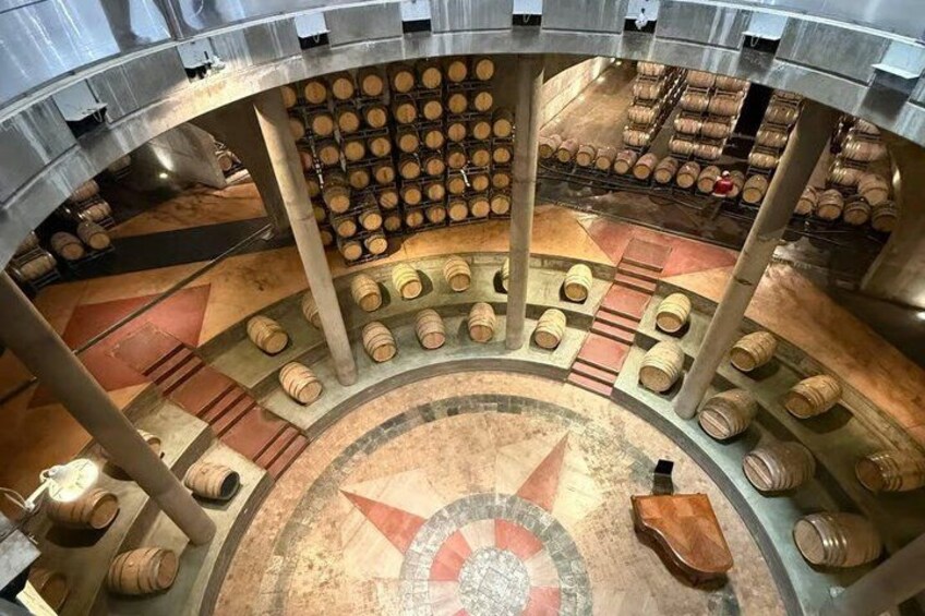 A circular room with wooden barrels arranged in tiers, a central circular design on the floor, and metal pillars supporting the structure. 4