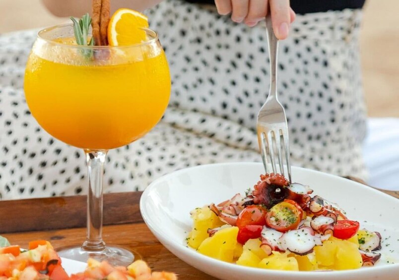 Un verre de jus d’orange avec une tige de cannelle et une tranche d’orange, une assiette de salade de fruits de mer avec des pommes de terre et des tomates, et une fourchette. 3