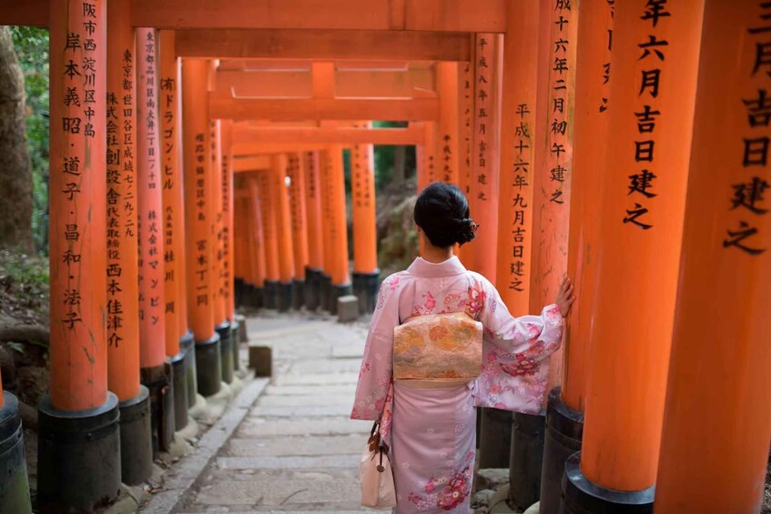 Una persona vestida con traje tradicional camina por un pasillo de torii anaranjados con inscripciones japonesas. 5
