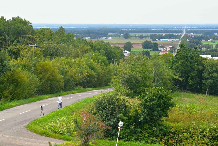 A winding road through a lush green landscape with a person walking and a camera on a tripod. 2