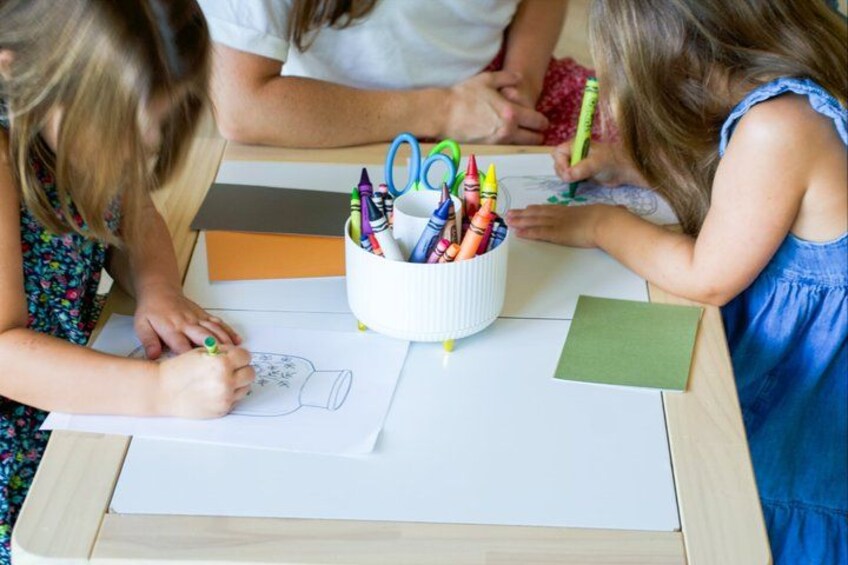 Children drawing with crayons on paper, a white container with crayons, and a green piece of paper on a table. 3