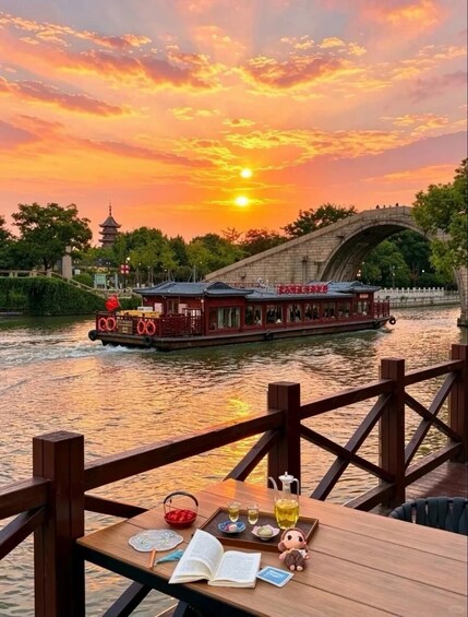 Un coucher de soleil sur une rivière, avec un bateau traditionnel, un pont en bois, et une table où sont posés des livres et des collations. 5