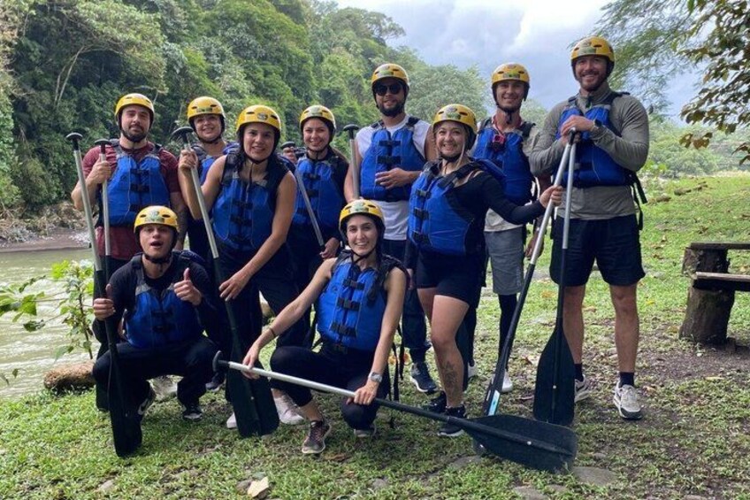 A group of people wearing helmets and life vests, holding paddles, standing on a grassy area near a river. 5