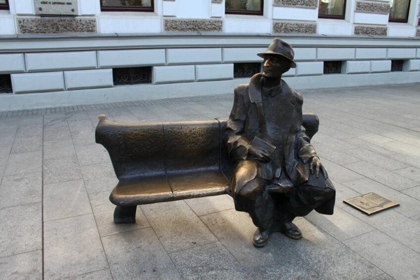 A bronze statue of a seated man with a hat and coat, holding a book, sitting on a bench. 2