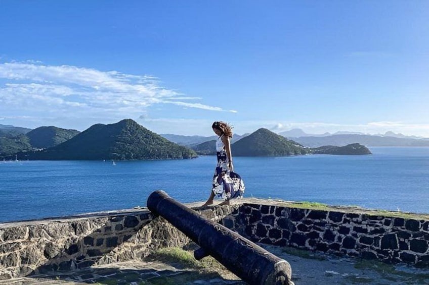 A person in a patterned dress walking along a stone wall by a body of water with mountains in the background. 2