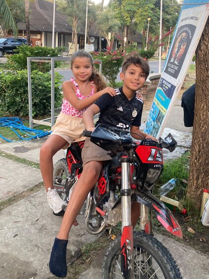 Two children on a dirt bike, a tree, and a signboard in the background. 2