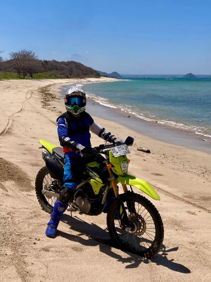 A person on a yellow dirt bike on a sandy beach with the ocean and hills in the background. 3