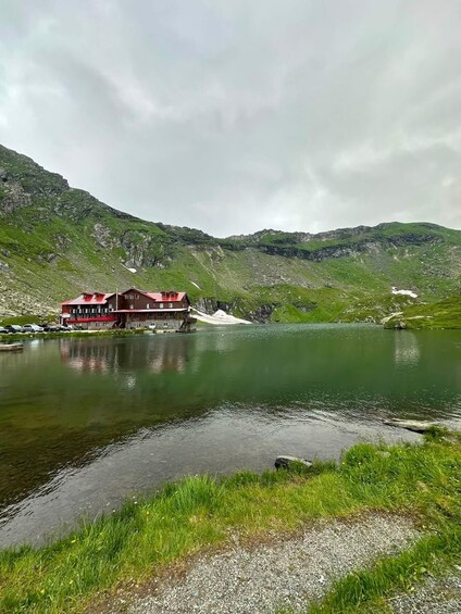 Ein Berggasthof an einem See mit rotem Dach, umgeben von grünen Hügeln. 1