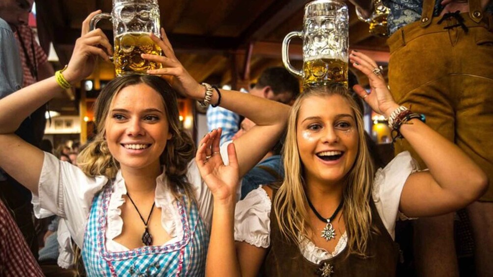 Two people holding beer mugs, wearing traditional attire, in a crowded indoor setting. 2