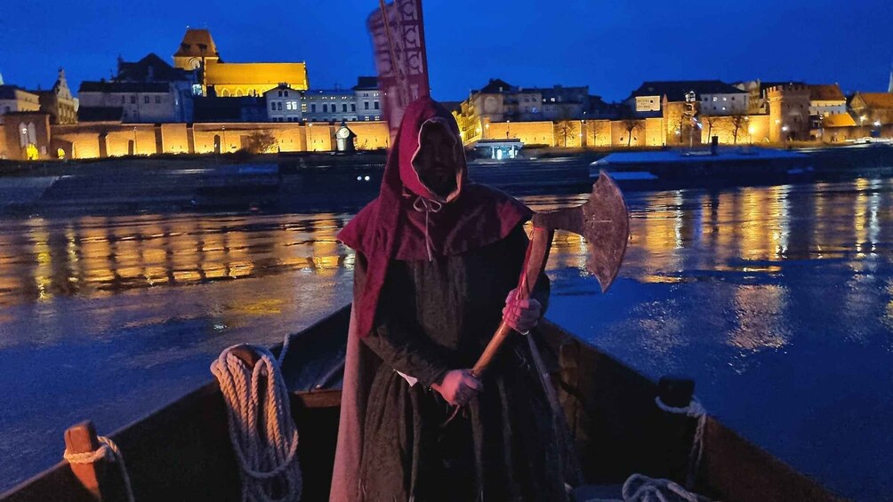 A person in a hooded cloak holding an axe on a boat with ropes, with a cityscape and buildings in the background. 1