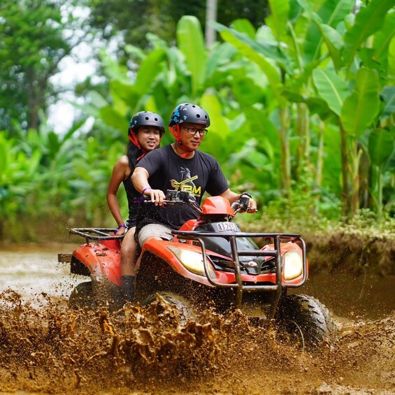 Zwei Personen fahren mit einem roten Geländefahrzeug durch einen schlammigen Bereich, im Hintergrund grüne Vegetation. 2