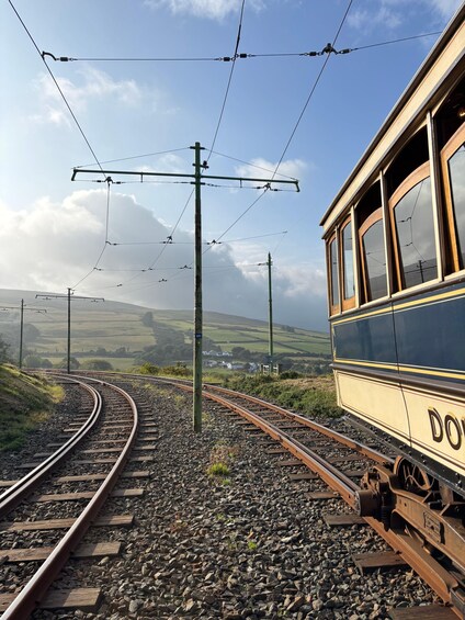 Un train d’époque circule sur une voie ferrée incurvée, surplombée par des lignes électriques aériennes, et est entouré d’un paysage rural composé de collines ondoyantes et de maisons dispersées. 5