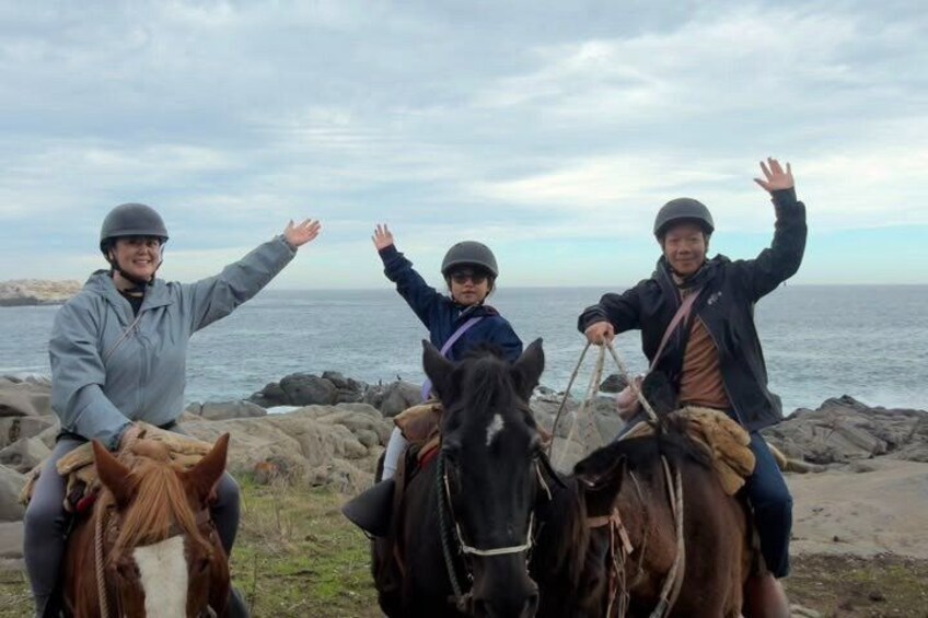Three people on horseback by a rocky shoreline with the ocean in the background. 3
