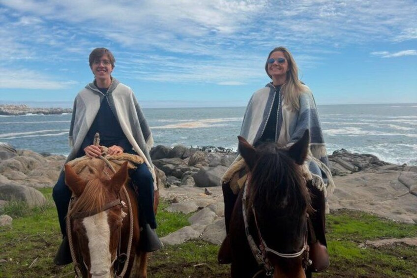 Two people on horseback by the ocean with rocky terrain and waves in the background. 4