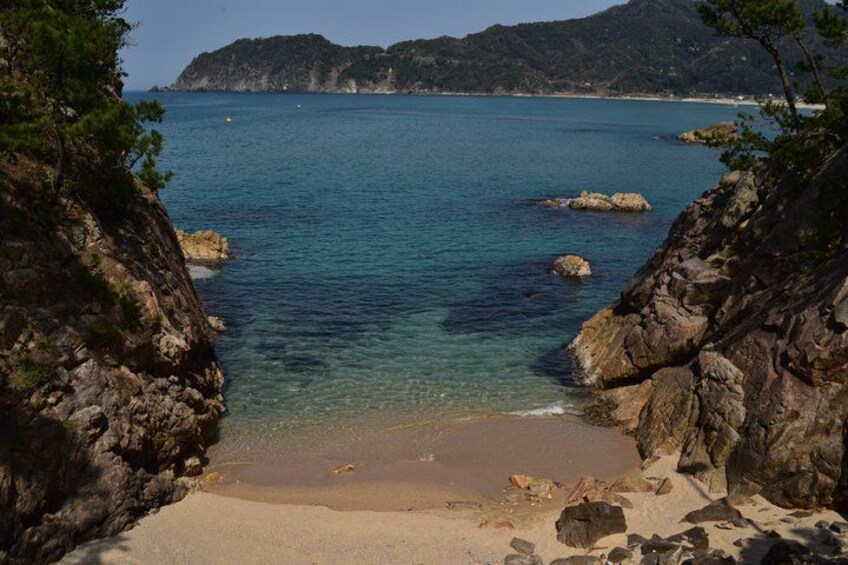 A coastal landscape with clear blue water, rocky shores, and a sandy beach. 1