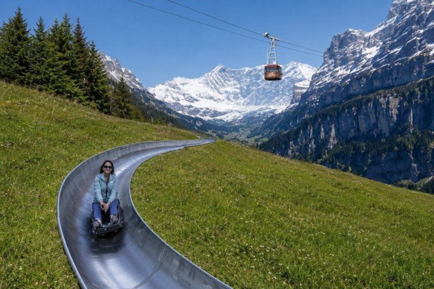 A person sliding down a metal slide in a mountainous area with a cable car in the background. 5