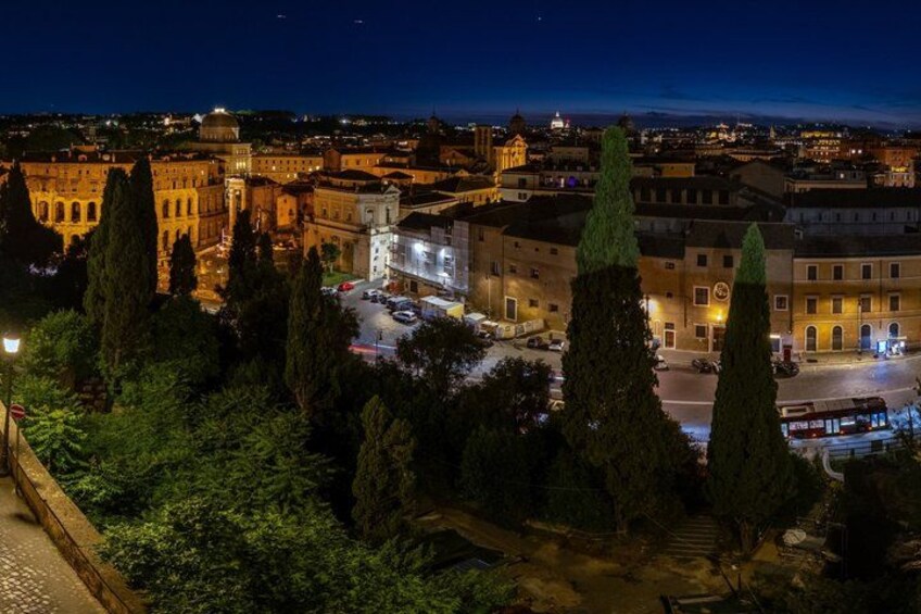 A nighttime cityscape with illuminated buildings, a prominent dome structure, and a central square with tall cypress trees. 3