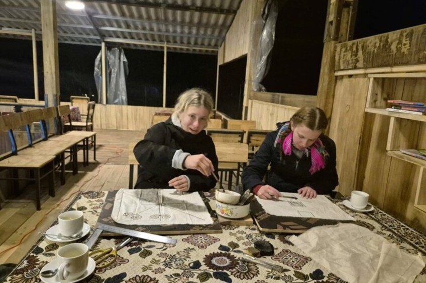 Two people at a wooden table with a tablecloth, working on a craft project. 5