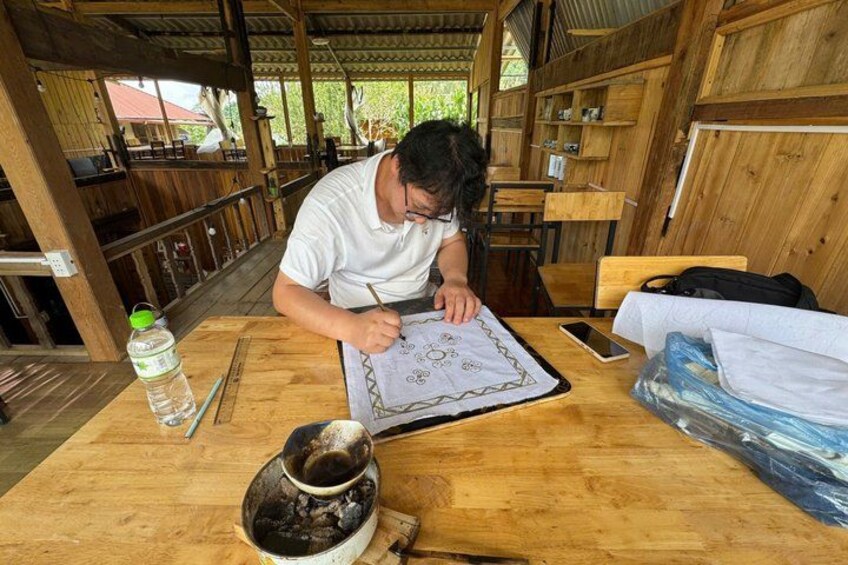 A person drawing on a piece of paper at a wooden table with a cup and a bottle nearby. 2