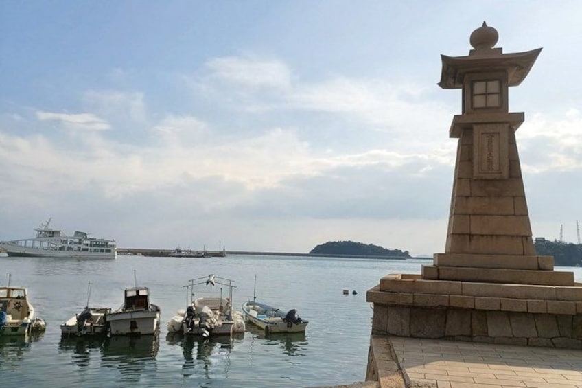 A calm harbor with several boats docked, a large ship in the distance, and a traditional stone monument on the right. 1