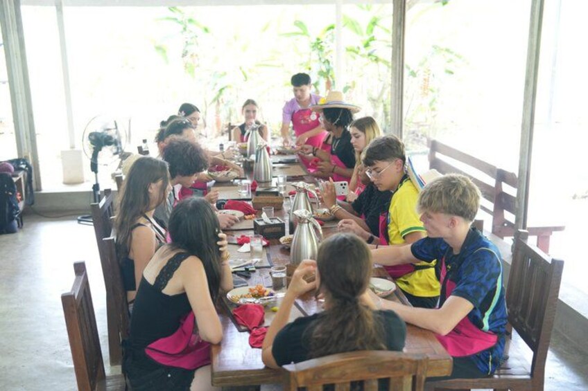 A group of people dining at a long table in a covered outdoor area with plants visible through the windows. 3