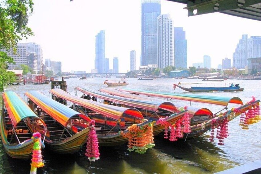 A river with colorful boats docked, a city skyline in the background, and a bridge overhead. 2