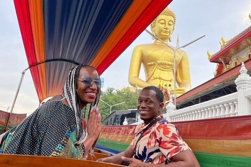 Two people in front of a large golden Buddha statue, with a colorful canopy and a temple in the background. 1
