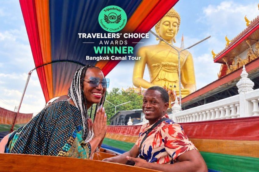 Two people in a boat with a large Buddha statue and colorful buildings in the background. 1