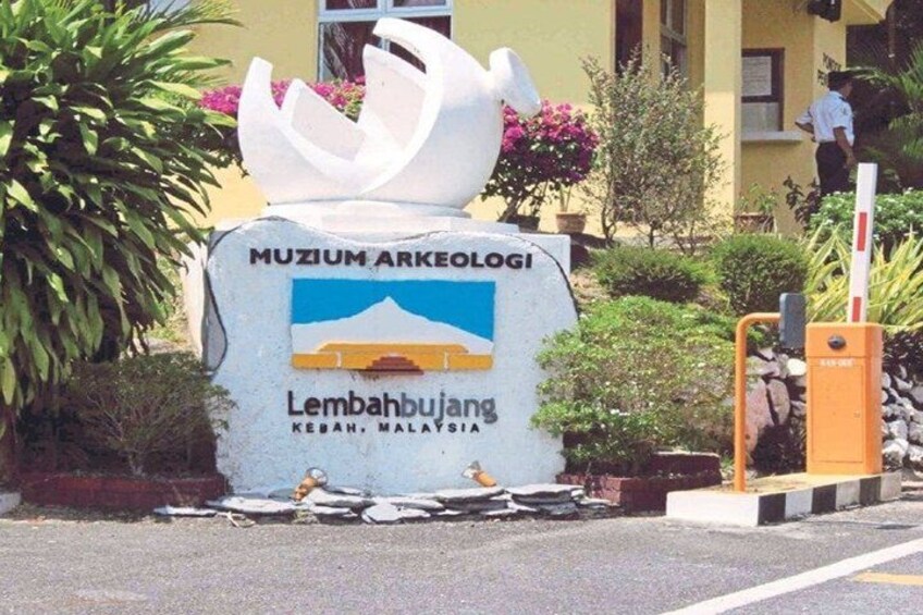 A sign for Muzium Arkeologi in Lembahbujang, Kebah, Malaysia, with a stylized white sculpture and a blue and yellow logo. 1