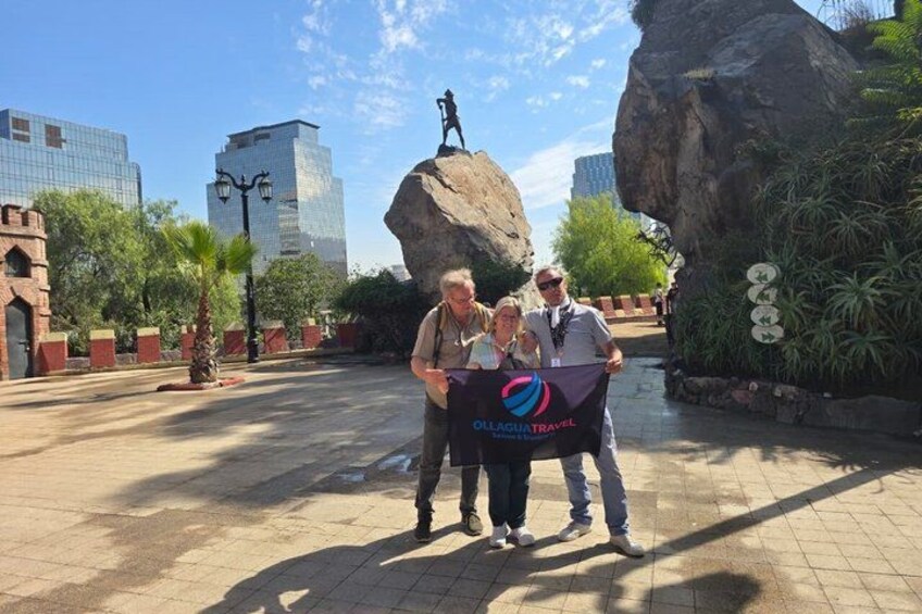 A group of people standing in front of a large rock formation with a statue on top, in an urban plaza with buildings and trees in the background. 5