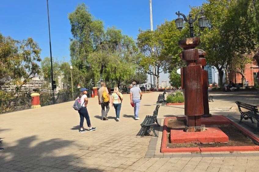 A paved walkway with benches, a large wooden sculpture, and trees in the background. 4