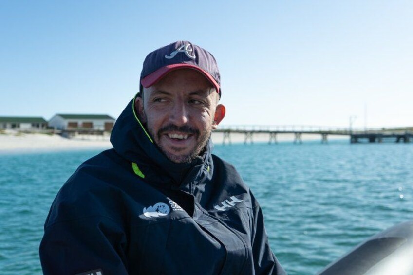 A person wearing a dark jacket and a cap, standing near a body of water with a pier in the background. 4