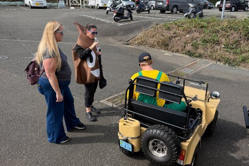 A person in a yellow vehicle with a license plate, another person in a brown jacket, and a third person in a gray shirt. 2
