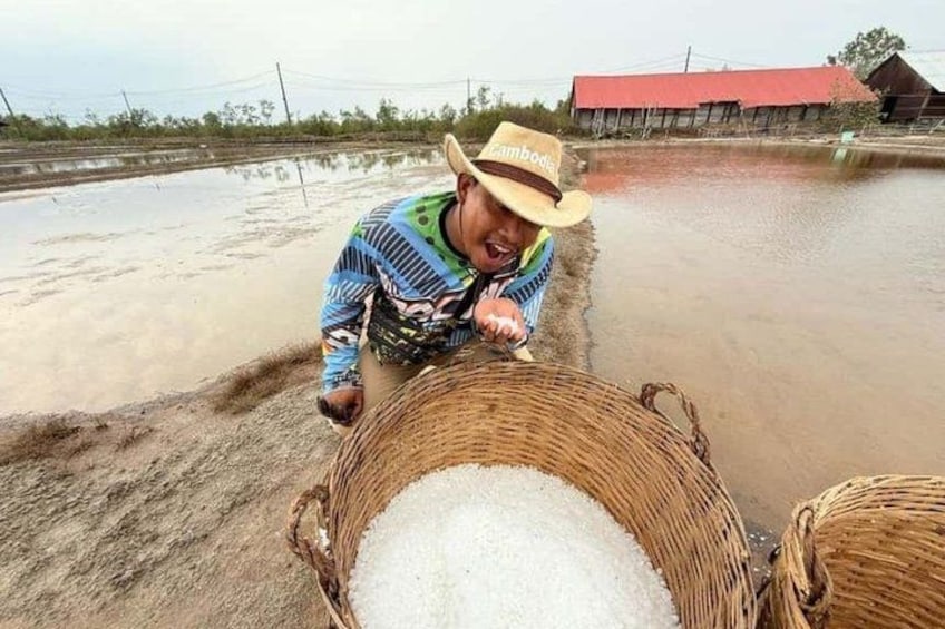 A person in a hat and striped shirt handling a basket of white material, possibly salt, in a rural setting with a red building in the background. 1