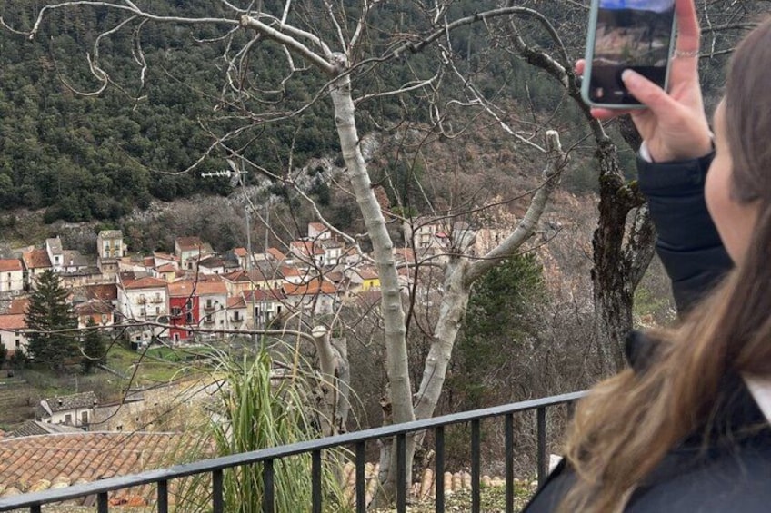 A person taking a photo of a town with red-roofed buildings and a hill in the background. 2