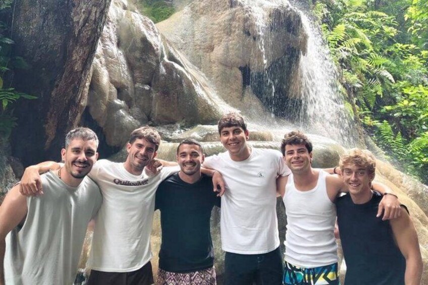 A group of people standing in front of a waterfall with greenery around. 4