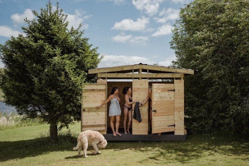 A wooden structure with two people standing at the entrance, a dog nearby, and a tree in the background. 4