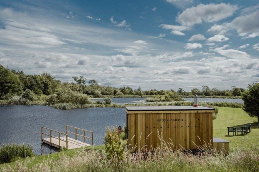 A wooden structure by a lake with a dock, surrounded by greenery and a clear sky. 1