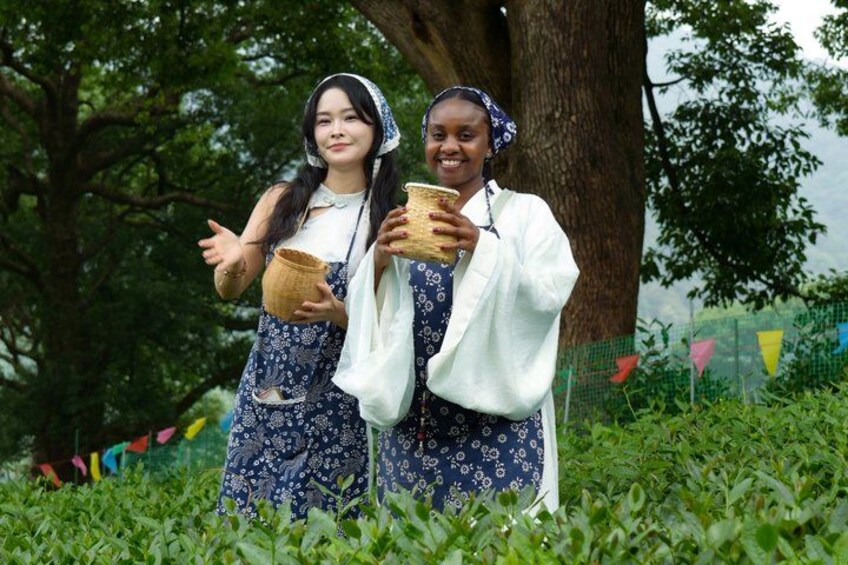 Two people in patterned dresses holding woven baskets in a grassy area with trees and a fence in the background. 1