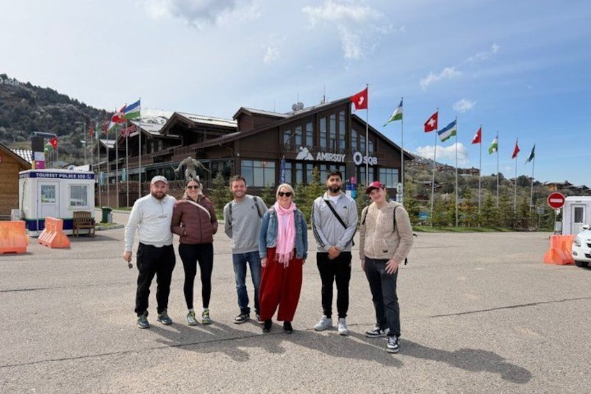 A group of people standing in front of a building with flags, a tourist police booth, and a mountain in the background. 3